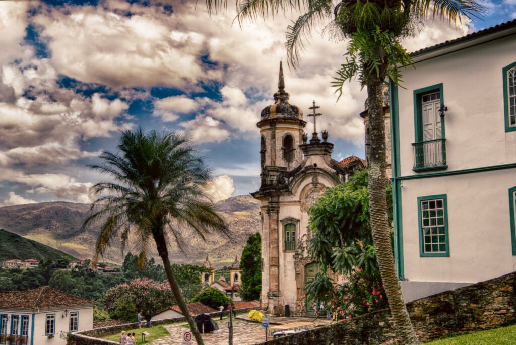 Igreja barroca colonial com torre sineira e cruz ao lado de casarão com janelas verdes, palmeiras em primeiro plano e montanhas ao fundo, em Ouro Preto, Minas Gerais.