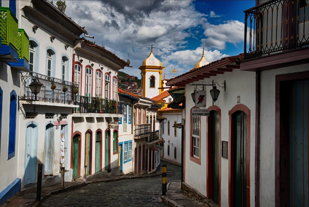 Rua de pedra com casarões coloniais coloridos e torres de igreja sob céu carregado em Ouro Preto.