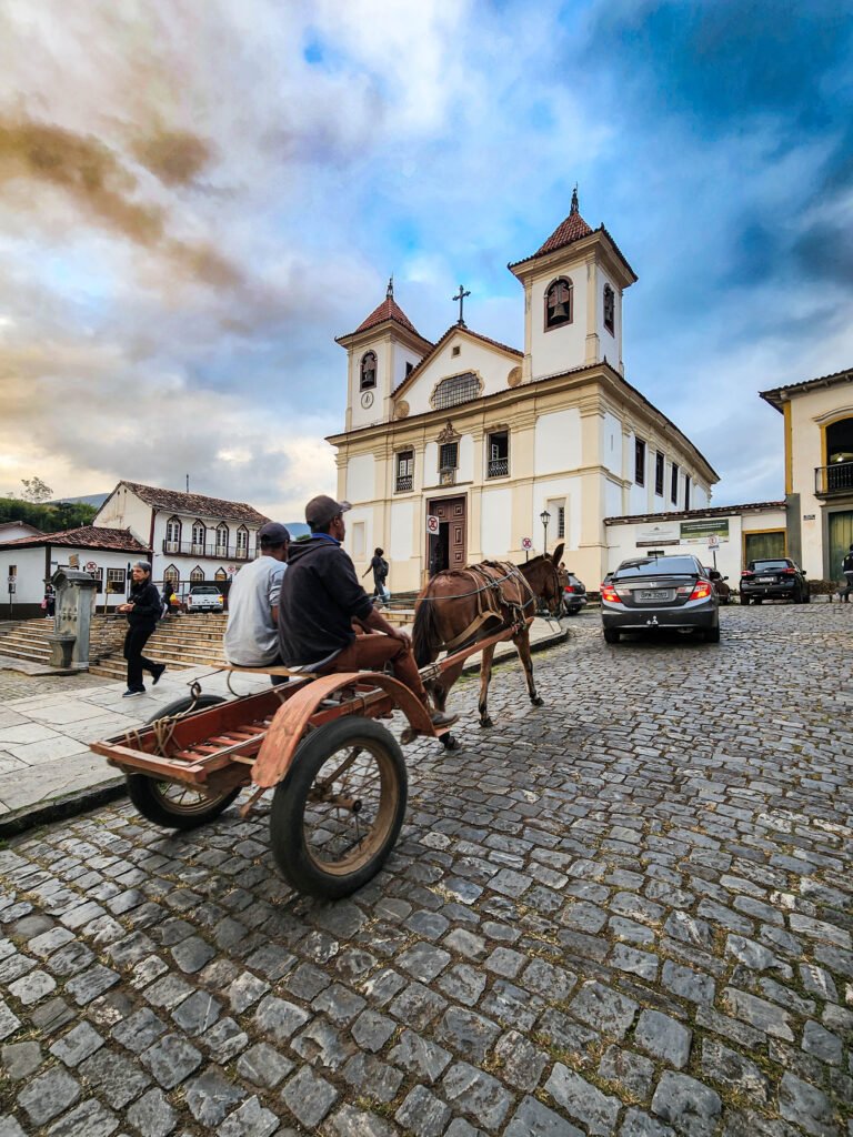 Carroça com duas pessoas diante de igreja, pedestres e automóveis na Praça da Sé, em Mariana.